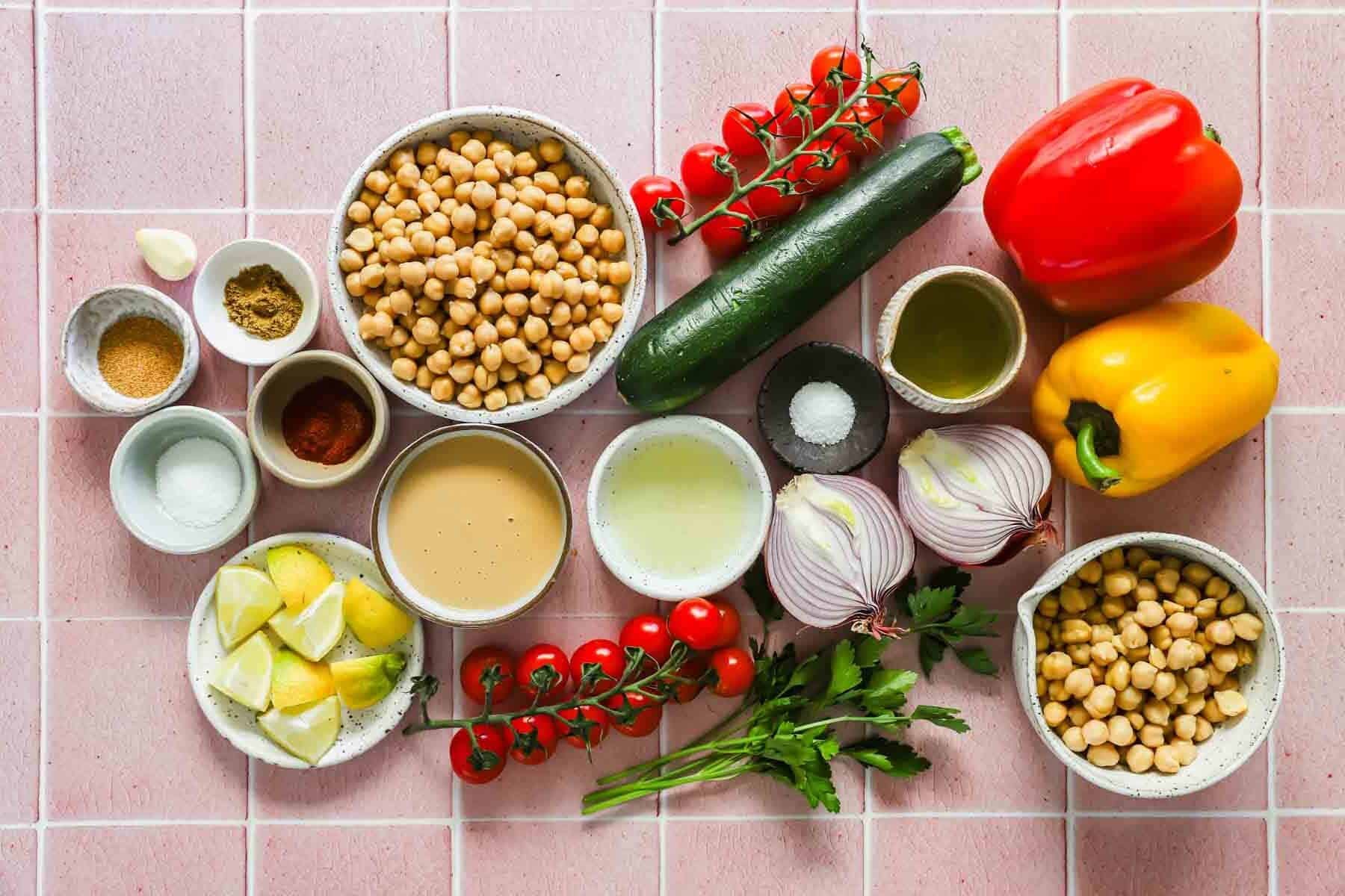 Assorted ingredients on a pink tiled surface including chickpeas, cherry tomatoes, zucchini, bell peppers, red onion, lemon wedges, spices, garlic, parsley, oil, and tahini.
