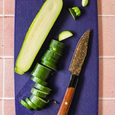 A partially sliced cucumber and a knife with a patterned blade rest on a dark cutting board atop a pink tiled surface.