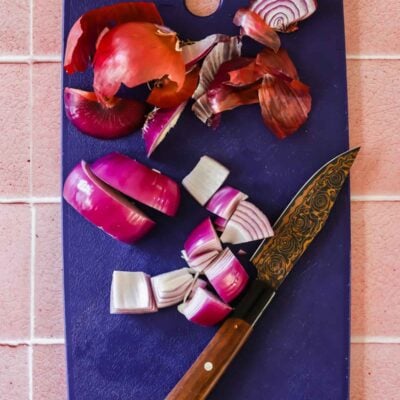 A cutting board with a partially chopped red onion, onion skin, and a knife on a pink tiled surface.