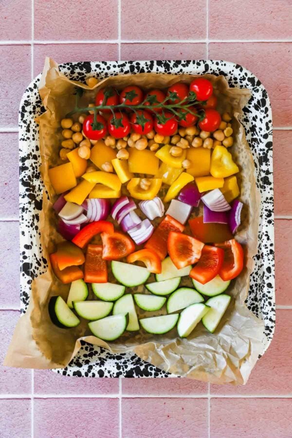A baking tray with parchment paper holds rows of grape tomatoes, chickpeas, yellow and red bell peppers, red onion, and sliced zucchini on a pink tiled surface.