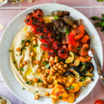 A bowl of hummus topped with roasted vegetables and chickpeas, surrounded by small bowls of ingredients and cherry tomatoes on a pink tile background.