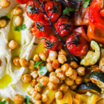 A bowl with hummus, roasted chickpeas, cherry tomatoes on the vine, zucchini, and yellow bell pepper, garnished with herbs and olive oil.