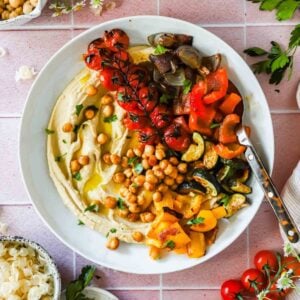 A plate of hummus topped with roasted chickpeas, cherry tomatoes on the vine, chopped red and yellow bell peppers, zucchini, and eggplant, served with a spoon.
