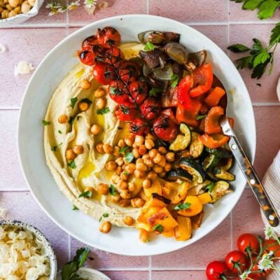 A plate of hummus topped with roasted chickpeas, cherry tomatoes on the vine, chopped red and yellow bell peppers, zucchini, and eggplant, served with a spoon.