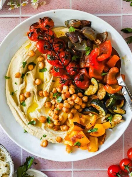 A plate of hummus topped with roasted chickpeas, cherry tomatoes on the vine, chopped red and yellow bell peppers, zucchini, and eggplant, served with a spoon.