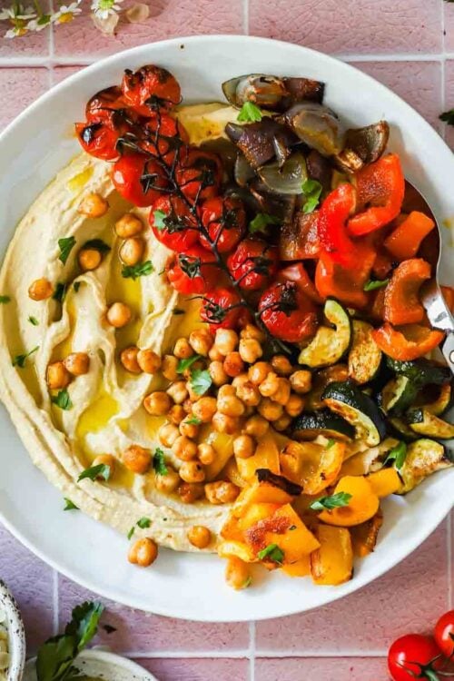 A plate of hummus topped with roasted chickpeas, cherry tomatoes on the vine, chopped red and yellow bell peppers, zucchini, and eggplant, served with a spoon.