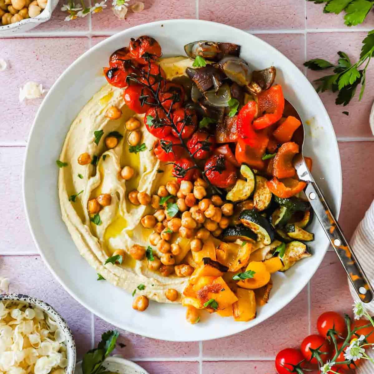 A plate of hummus topped with roasted chickpeas, cherry tomatoes on the vine, chopped red and yellow bell peppers, zucchini, and eggplant, served with a spoon.
