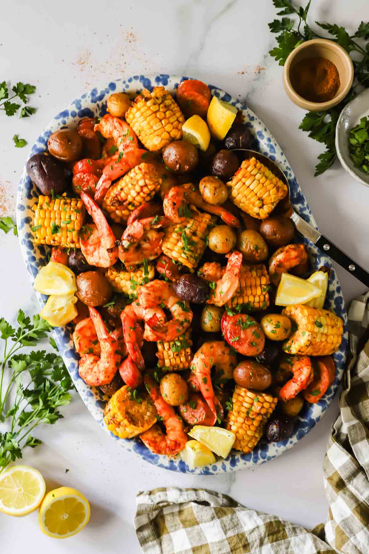 A platter of shrimp, corn on the cob pieces, baby potatoes, and lemon wedges garnished with herbs, set on a marble surface with parsley and other seasonings nearby.