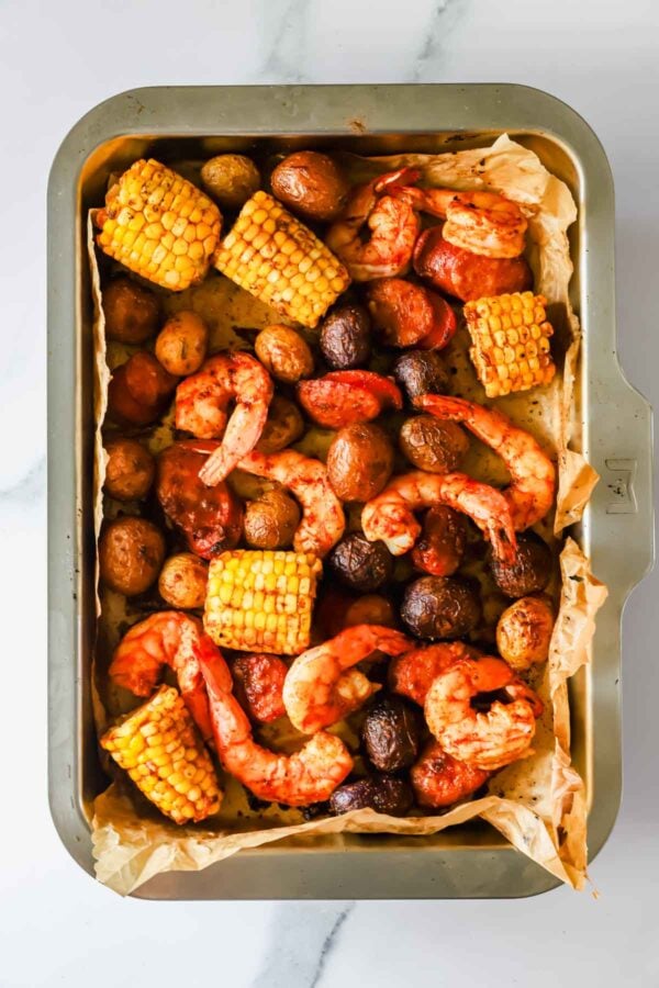 A baking tray lined with parchment paper holds roasted shrimp, baby potatoes, and corn on the cob pieces, all seasoned and cooked.