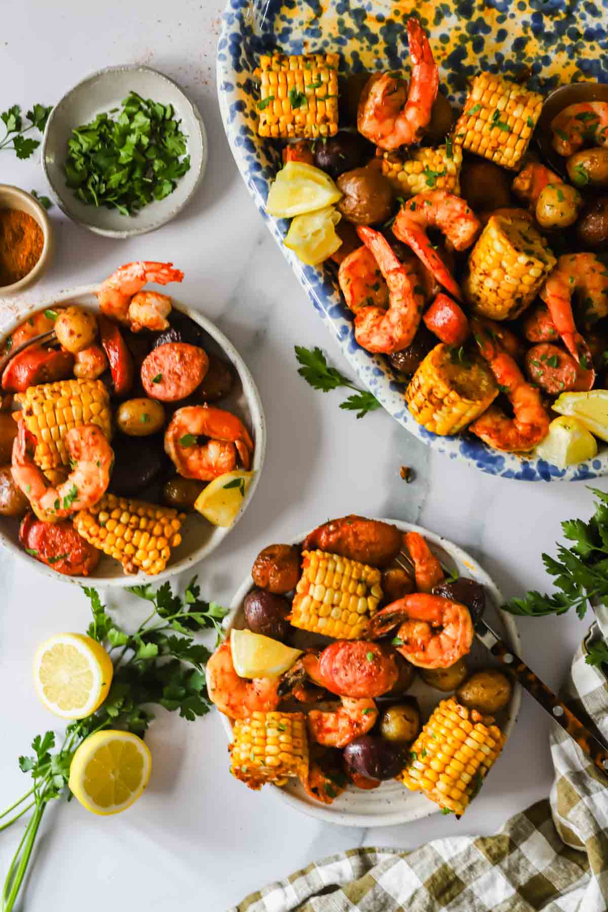 Plates and a large serving dish filled with shrimp, corn on the cob, potatoes, sausage, and lemon wedges, surrounded by fresh parsley and lemon slices on a white surface.