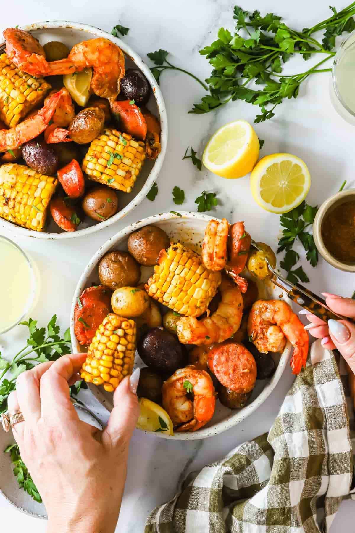 Two bowls filled with shrimp, corn on the cob, sausage, and potatoes, surrounded by fresh parsley, lemon halves, and a checkered cloth napkin. Hands are serving the food.