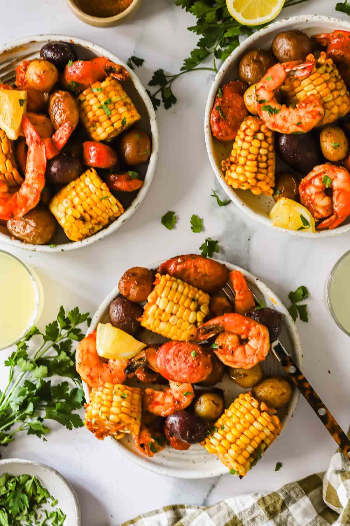 Three bowls containing shrimp, corn on the cob pieces, red potatoes, and lemon wedges, surrounded by parsley and drinks on a white surface.