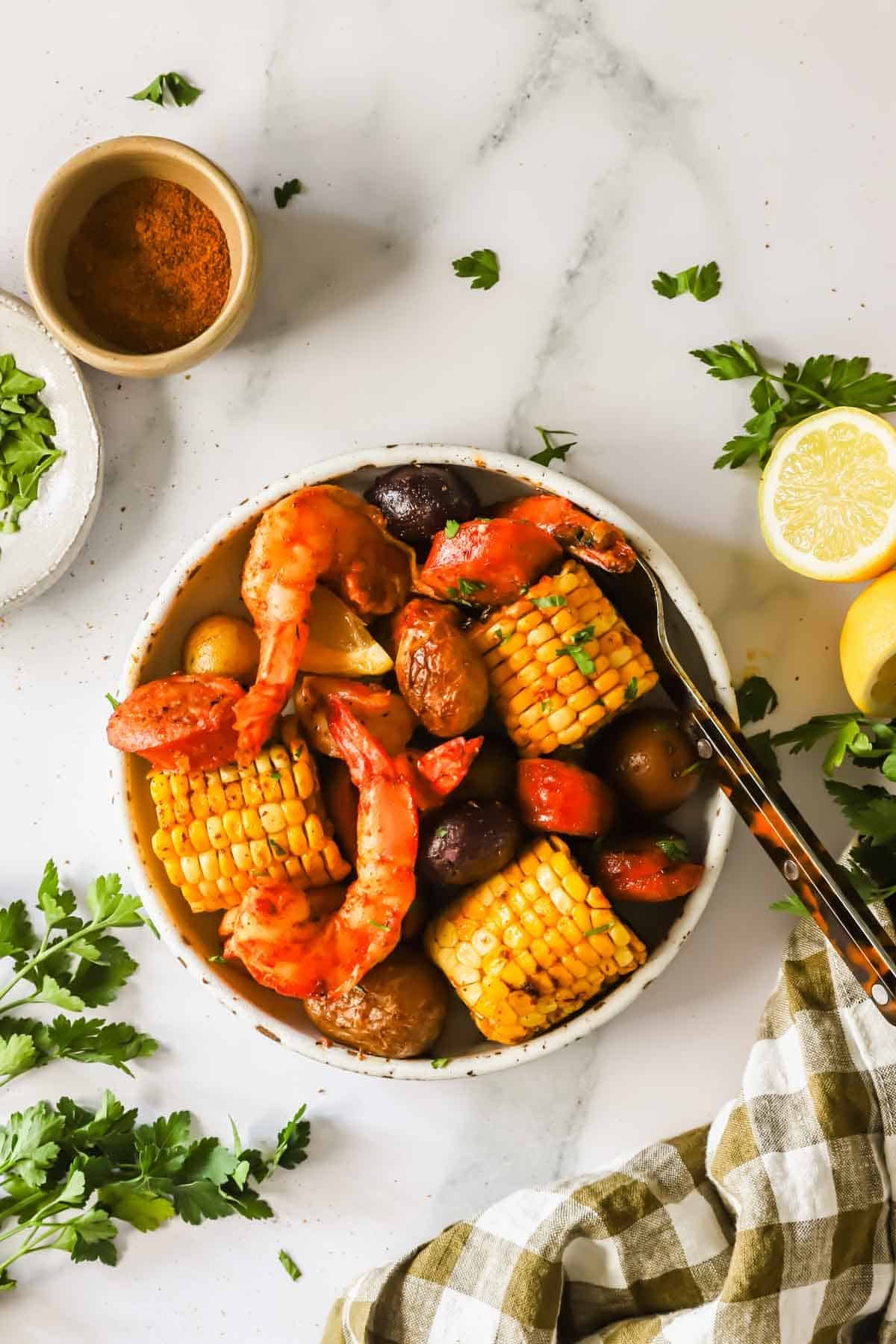 A bowl containing shrimp, crab legs, corn on the cob, potatoes, and sausage sits on a marble surface, surrounded by parsley, a lemon, seasoning, and a checkered cloth.