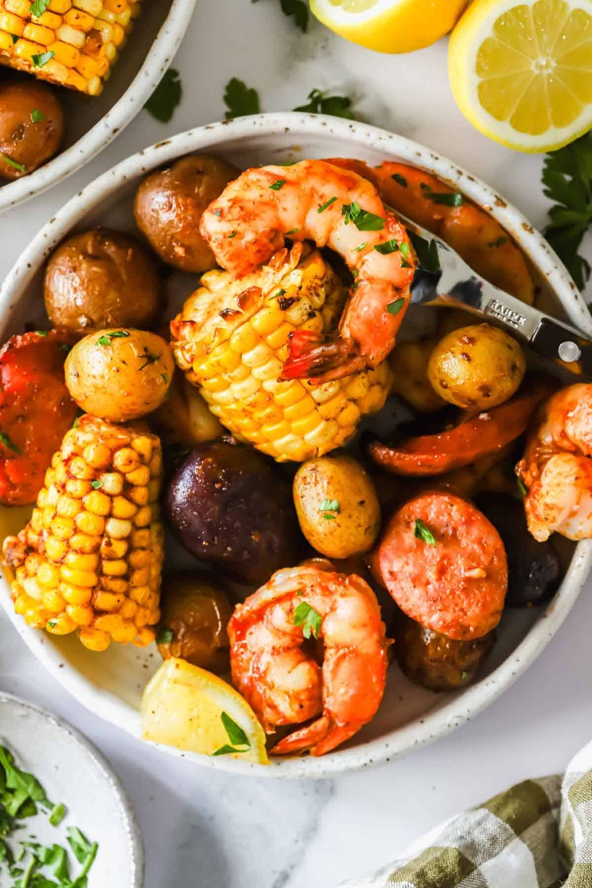 A bowl of shrimp, corn on the cob, baby potatoes, and sausage slices garnished with chopped herbs, with lemon halves and more food visible in the background.