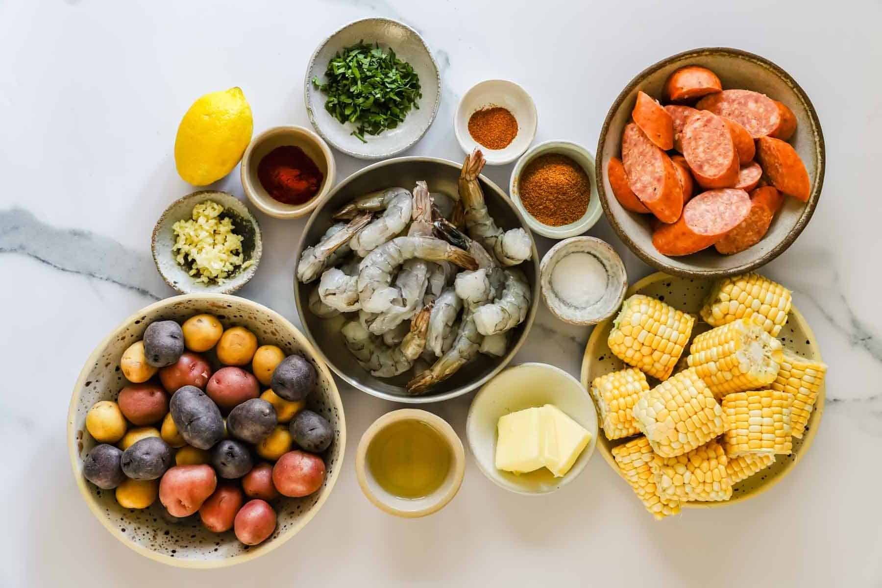 Assorted ingredients for a seafood boil, including raw shrimp, sliced sausage, corn, potatoes, butter, lemon, garlic, herbs, spices, and oil, arranged in bowls on a white surface.