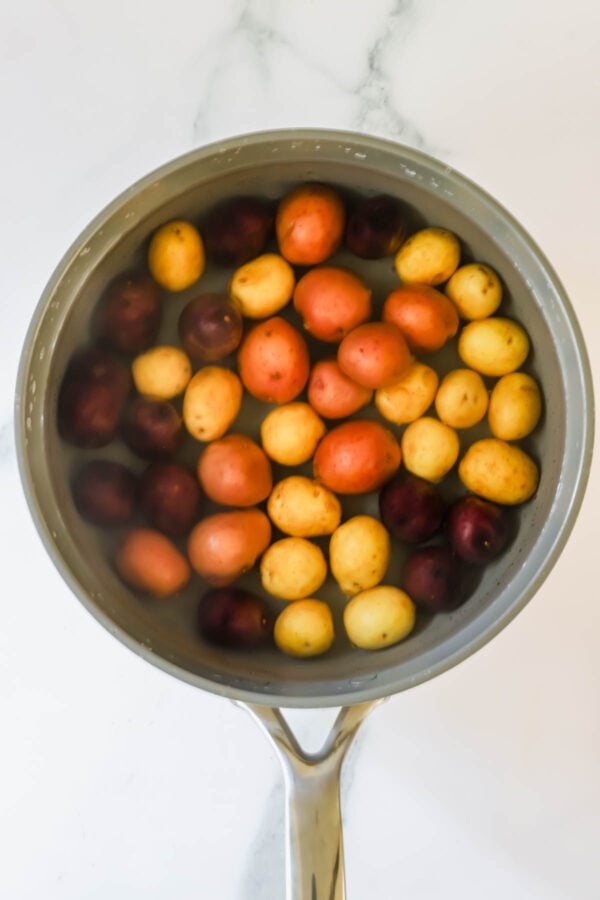 A saucepan filled with water and mixed small potatoes—red, yellow, and purple varieties—on a white marble surface.