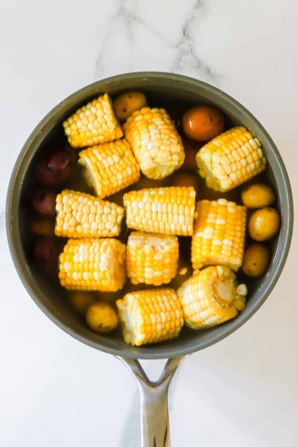 A saucepan filled with water contains cut corn on the cob and small whole potatoes, placed on a white marble surface.