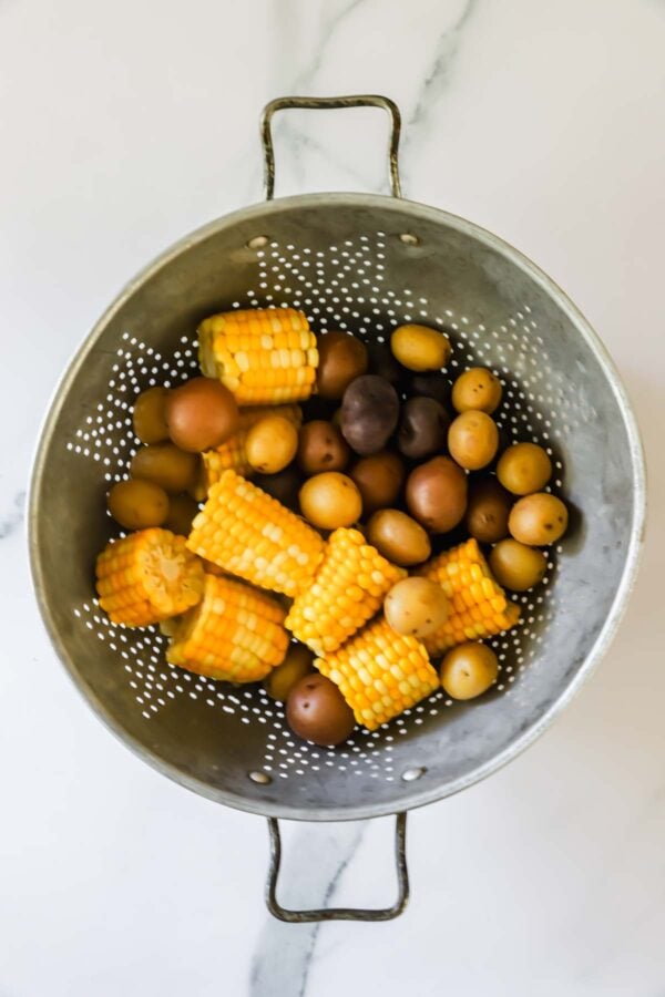 A metal colander containing halved corn on the cob and mixed small potatoes on a white surface.
