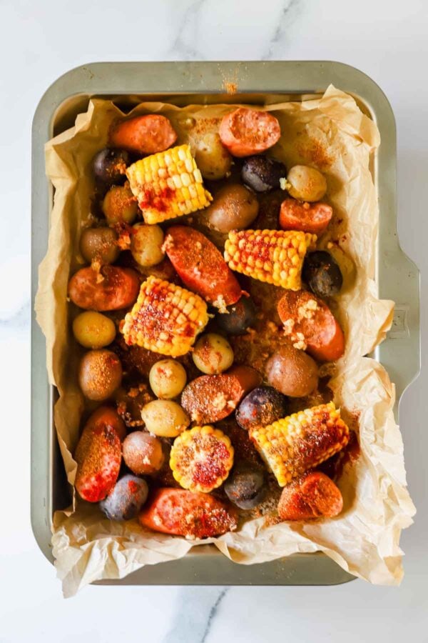 A baking tray lined with parchment paper holds roasted corn on the cob pieces, small potatoes, and sliced sausage, seasoned with spices.
