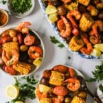 Plates of shrimp boil with corn on the cob, potatoes, sausage, shrimp, lemon wedges, and parsley, arranged on a white surface.