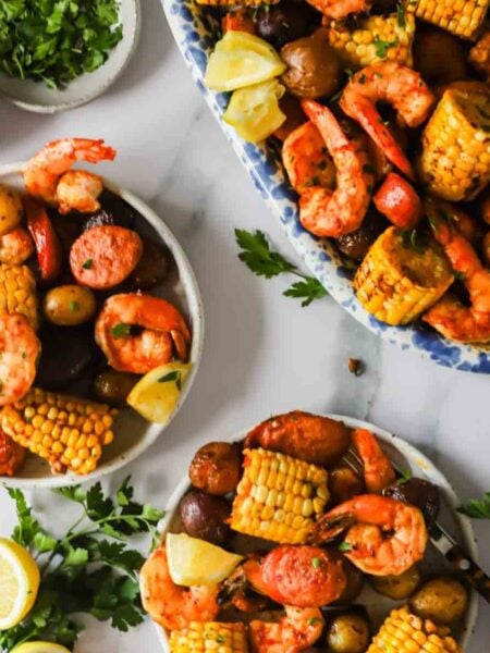Plates of shrimp boil with corn on the cob, potatoes, sausage, shrimp, lemon wedges, and parsley, arranged on a white surface.
