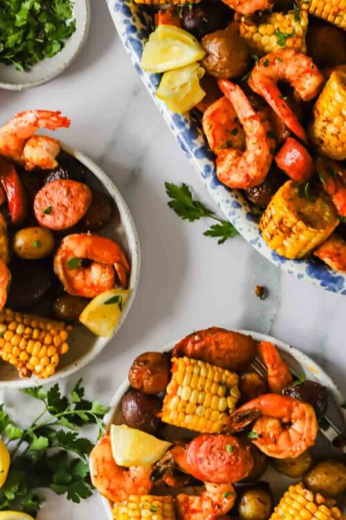 Plates of shrimp boil with corn on the cob, potatoes, sausage, shrimp, lemon wedges, and parsley, arranged on a white surface.