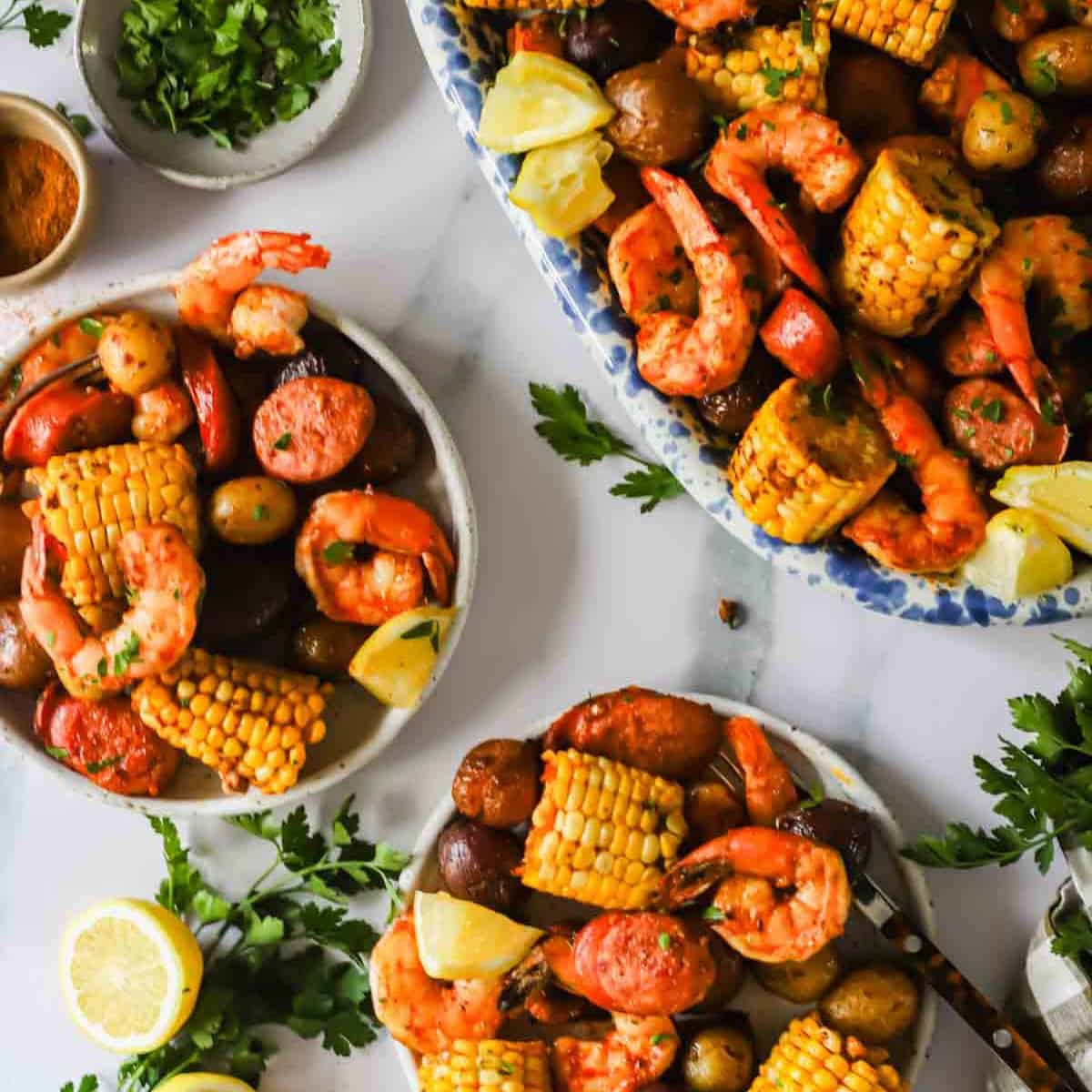 Plates of shrimp boil with corn on the cob, potatoes, sausage, shrimp, lemon wedges, and parsley, arranged on a white surface.