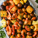 A platter of sheet pan shrimp boil with shrimp, corn on the cob, potatoes, lemon wedges, and herbs, displayed on a patterned dish.