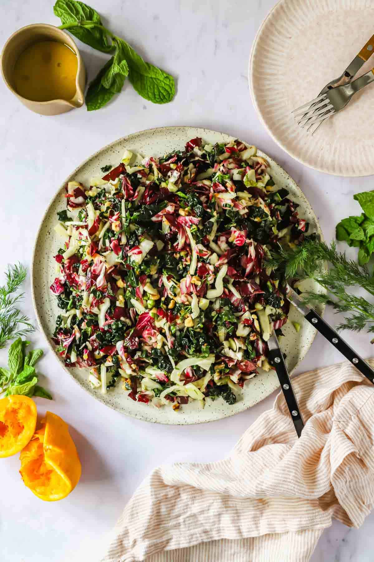 A large plate of chopped radicchio fennel salad with leafy greens, herbs, and seeds sits on a table with serving utensils, fresh herbs, halved orange, and a bowl of dressing nearby.