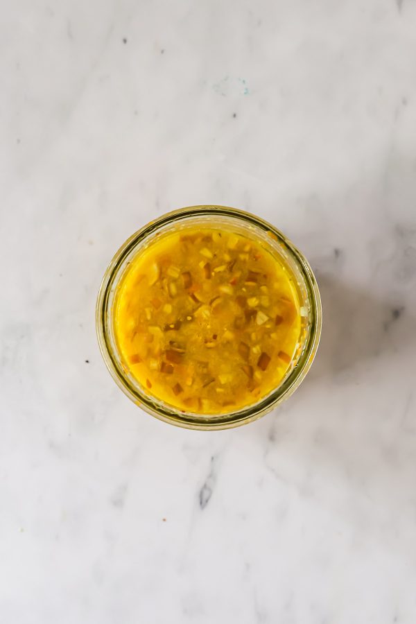 A glass jar filled with a yellow liquid containing finely chopped ingredients, placed on a light marble surface.