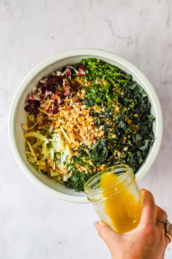 A hand pours yellow salad dressing from a jar onto a bowl of chopped vegetables and nuts, including greens, radicchio, and herbs, on a marble surface.