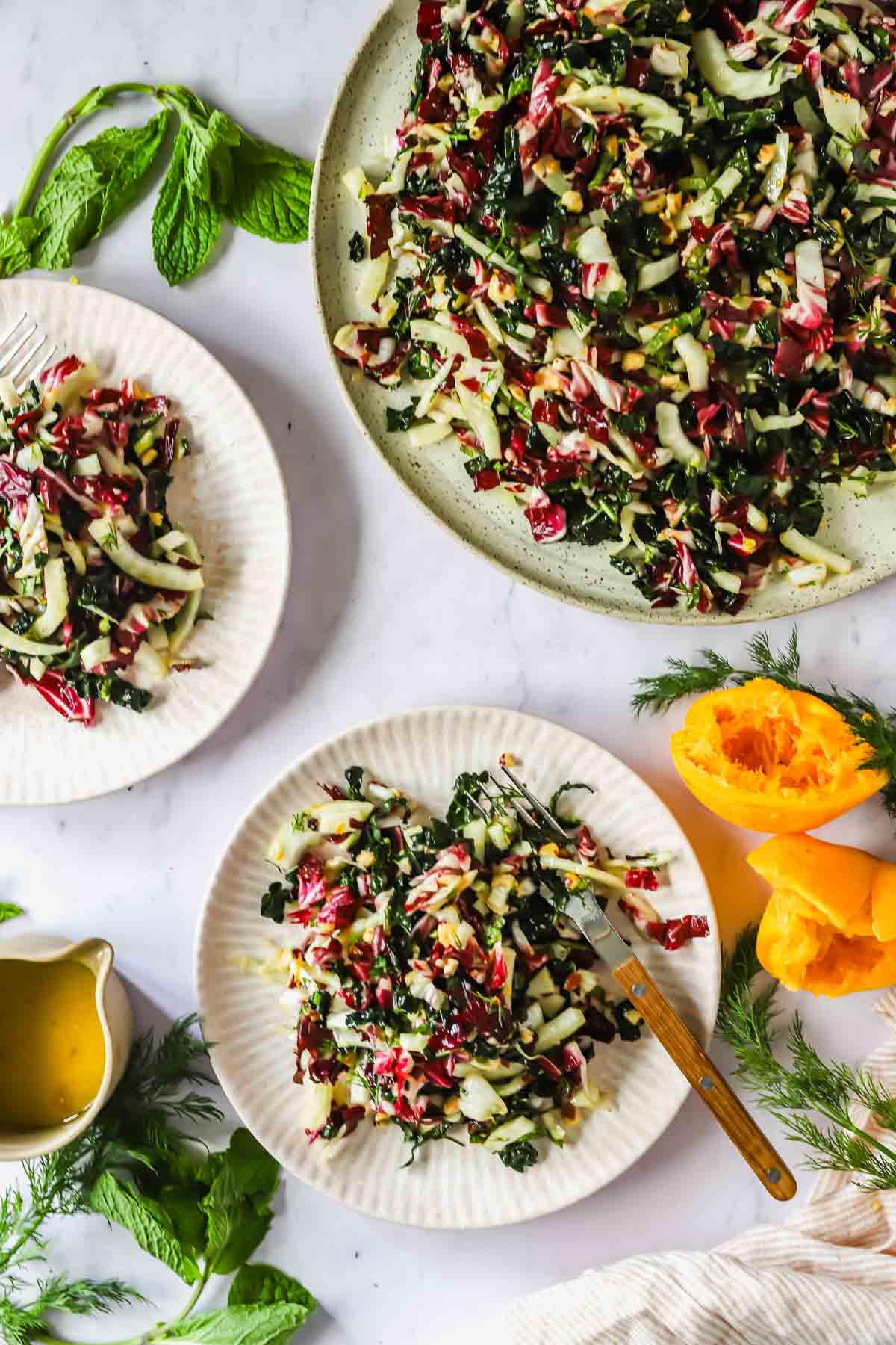 Three plates of leafy green salad with shredded vegetables on a white surface, accompanied by a halved yellow squash, fresh herbs, and a small pitcher of dressing.