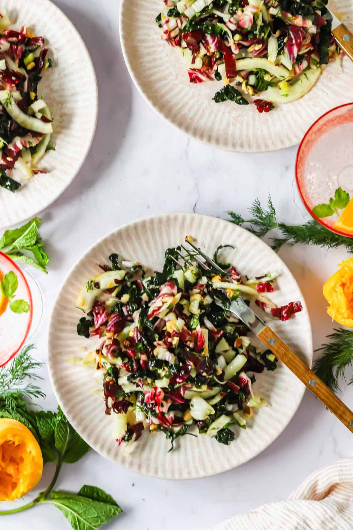 Four plates of mixed green salad with shredded kale, radicchio, and fennel are arranged on a white surface near fresh herbs, an empty glass, and squeezed citrus halves.