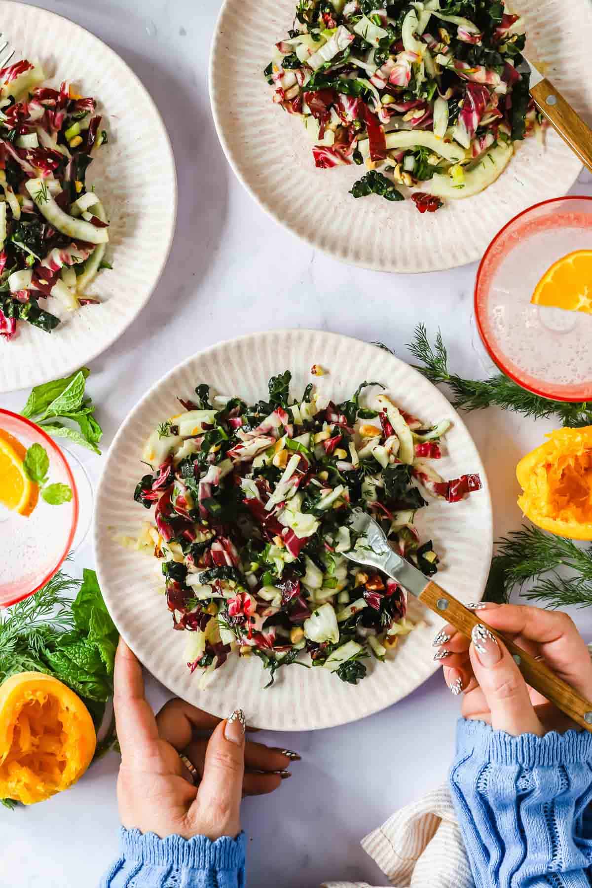 A person holds a fork and plate of mixed leafy salad with red and green vegetables, next to orange halves, herbs, and drinks with orange slices on a white table.