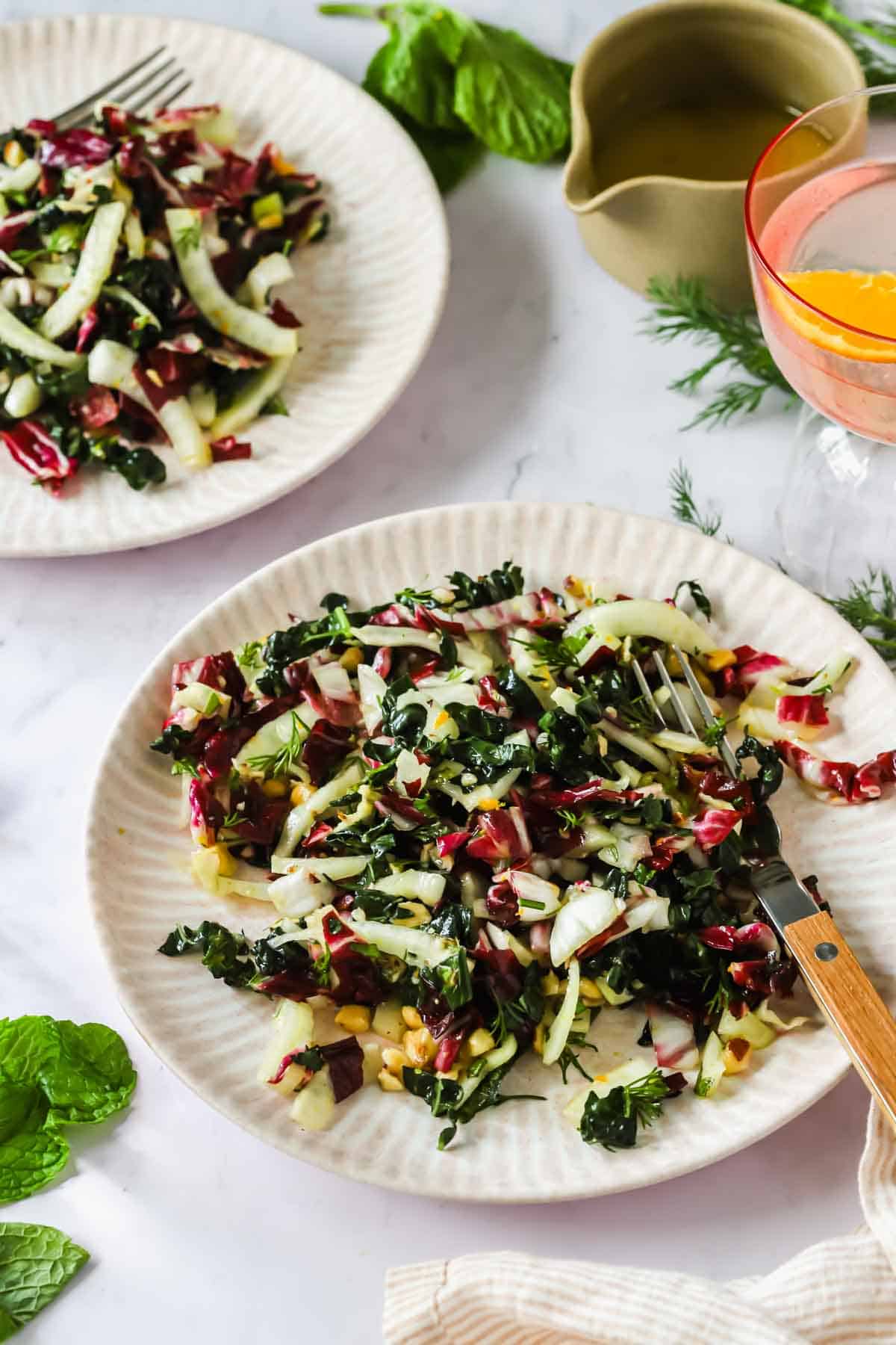 Two plates of mixed green salad with kale, radicchio, and fennel, served with a fork. A jug of dressing and a glass of ros&eacute; are nearby on a white surface.