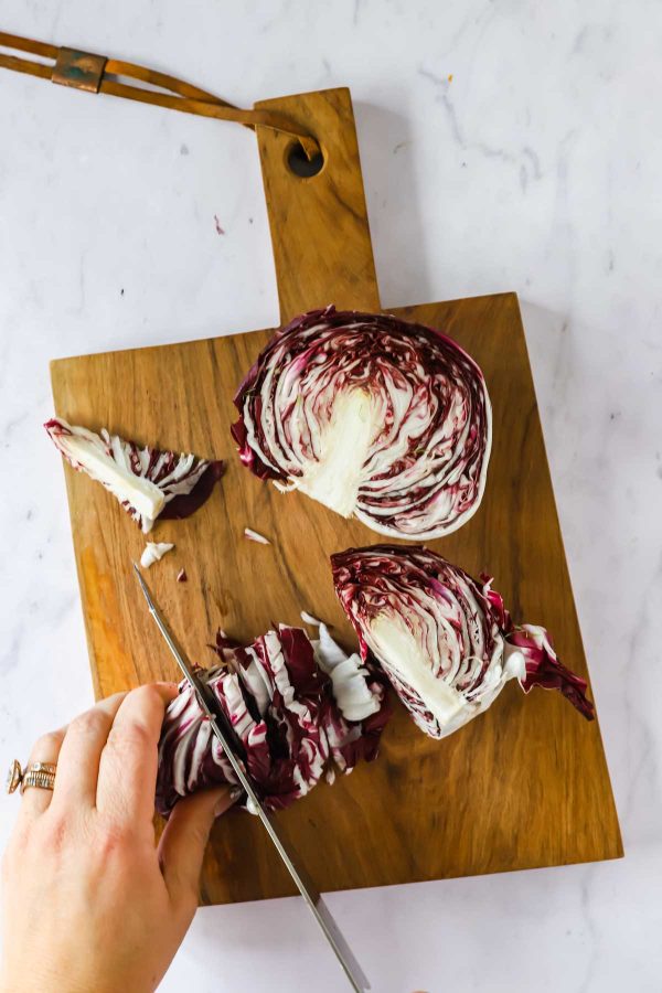 A hand slices a head of radicchio on a wooden cutting board with a knife, on a white countertop.