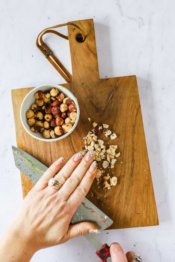 A hand using a knife to crush hazelnuts on a wooden cutting board, with a bowl of whole hazelnuts nearby.