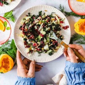 A person holds a plate of chopped salad with greens and radicchio, using a fork. Fresh herbs, citrus halves, and drinks are on the table.