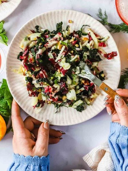 A person holds a plate of chopped salad with greens and radicchio, using a fork. Fresh herbs, citrus halves, and drinks are on the table.