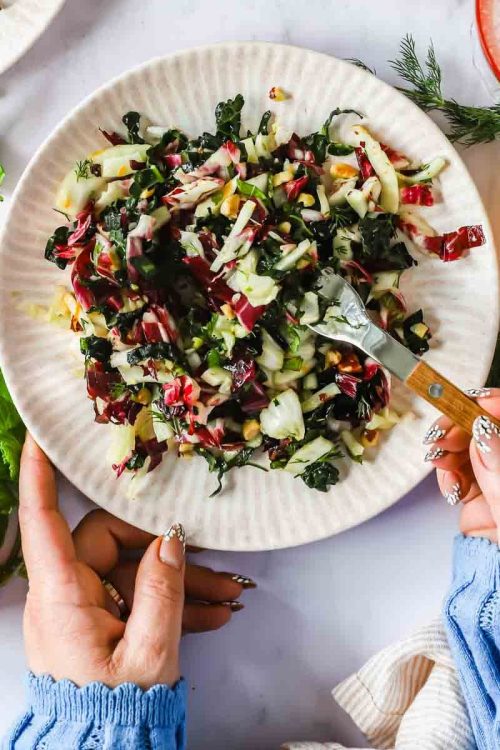 A person holds a plate of chopped salad with greens and radicchio, using a fork. Fresh herbs, citrus halves, and drinks are on the table.