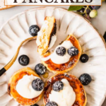 A plate with four cottage cheese pancakes topped with whipped cream, blueberries, and powdered sugar, with a fork and additional berries on the side.