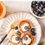 A plate with three Ukrainian syrniki pancakes topped with cream and blueberries, a fork, a jar of honey, and a bowl of blueberries beside it.