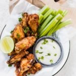 Plate of air-fried chicken wings with celery sticks, a lime wedge, and a bowl of dipping sauce garnished with chopped green onions. Text at the top reads "The Best Air Fryer Chicken Wings.