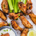 Air fried chicken wings arranged on parchment with celery sticks, a bowl of ranch dip, lime wedges, and green onion garnish.