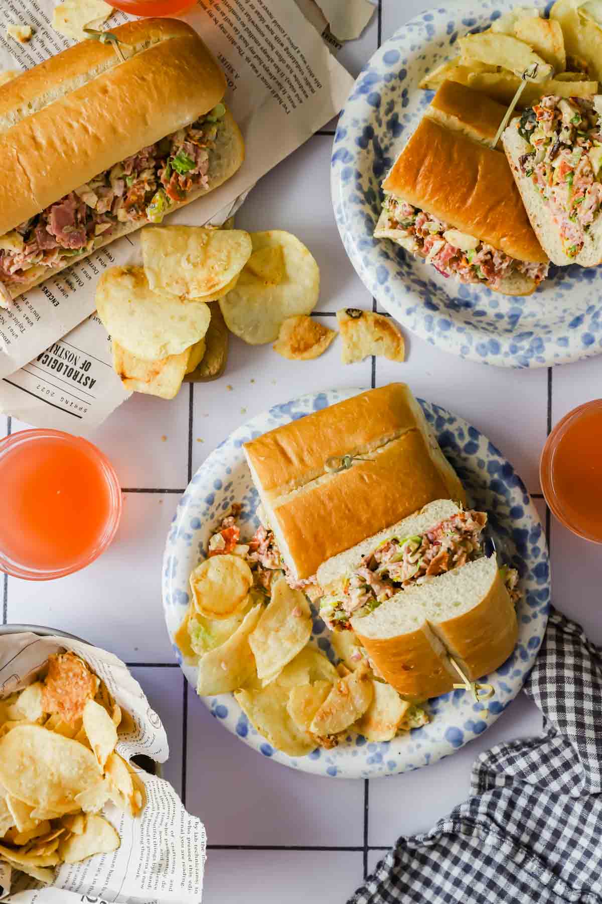 Three plates with sub sandwiches and potato chips, two glasses of orange drink, and extra chips on a white tiled surface with a checkered cloth and newspaper.