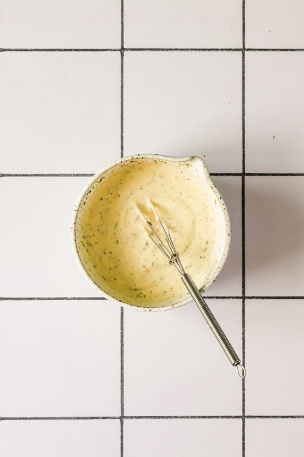 A small whisk rests in a bowl of creamy, speckled sauce on a white tiled surface.