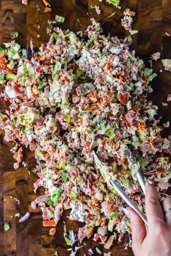 Chopped salad with assorted vegetables, meats, and dressing being mixed with tongs on a wooden surface.