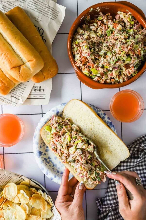 A person fills a sandwich bun with tuna salad from a bowl; two drinks and a plate of potato chips are on the table, along with extra sandwich buns.