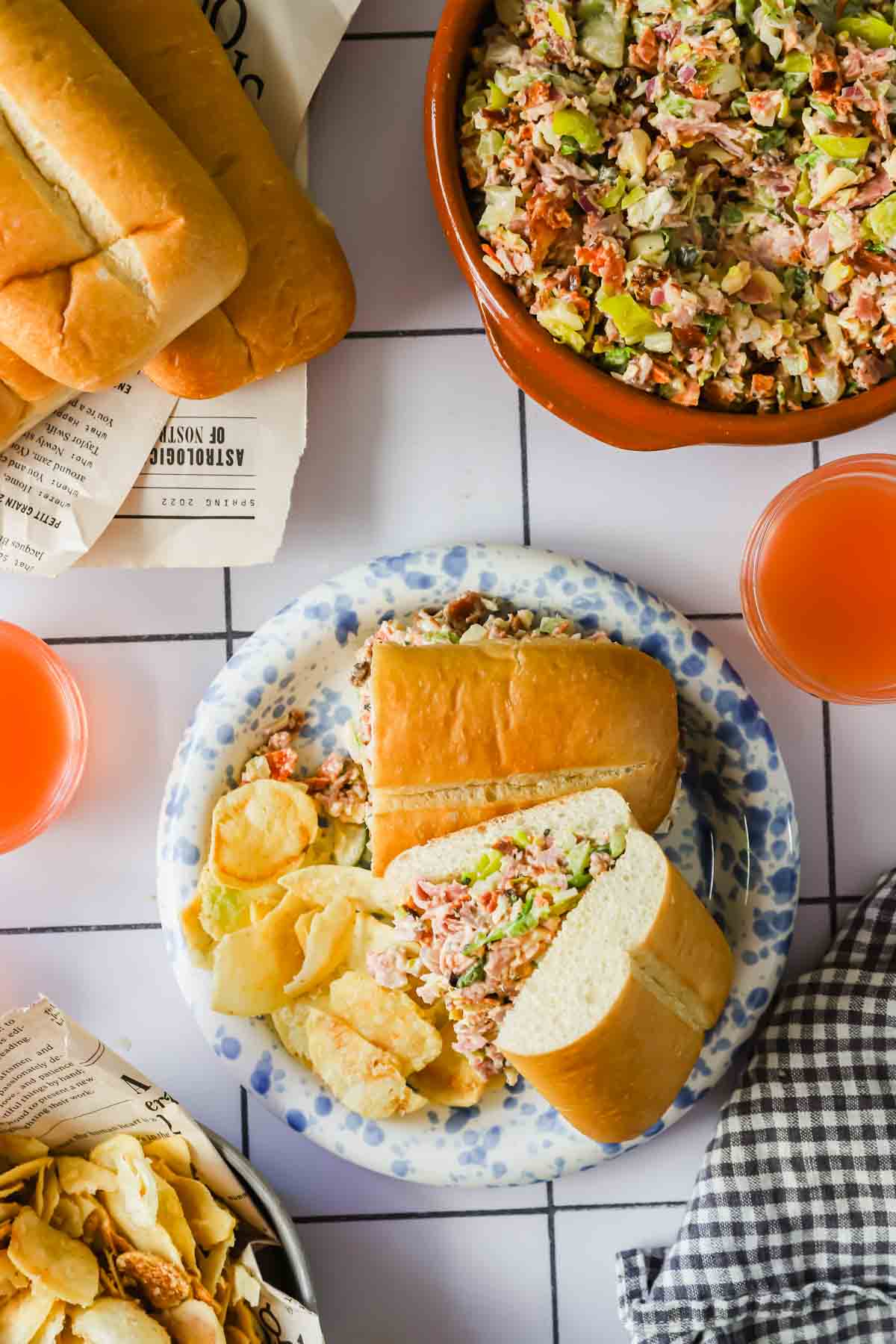 A plate with a sub sandwich filled with a salad mixture and potato chips, surrounded by a bowl of salad, drinks, bread rolls, and a napkin on a tiled table.