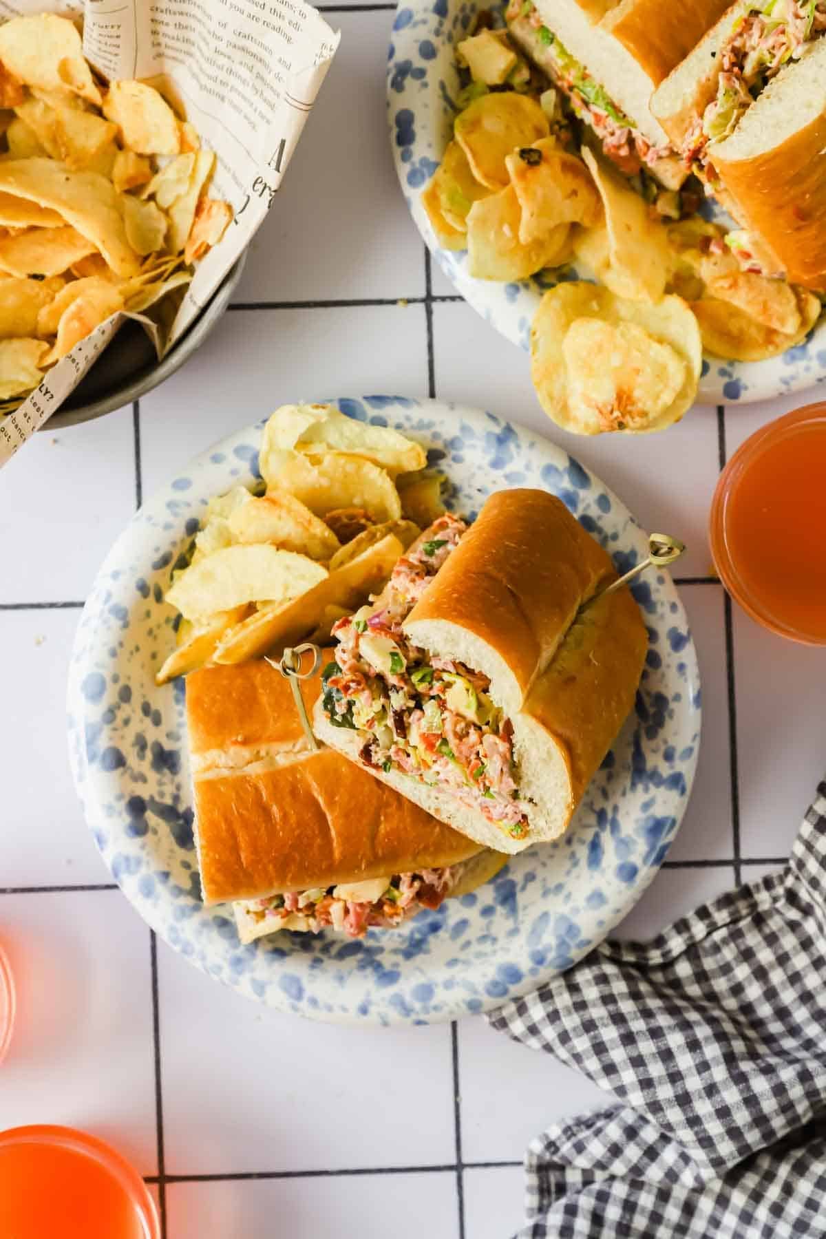 Two plates with sub sandwiches filled with deli meat and vegetables, each served with potato chips. Nearby are glasses of orange drink, a basket of chips, and a checkered napkin.